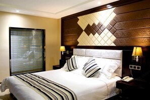 Quarto de hotel com uma cama grande, uma mesa de cabeceira com abajur, um telefone e uma janela com persianas.
