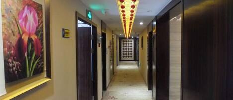 A hotel corridor with doors on both sides, a patterned carpet, and a ceiling with hanging lights.