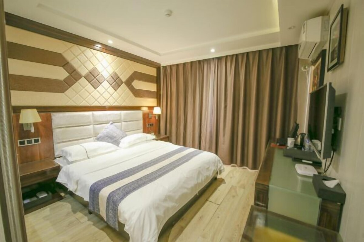 Une chambre d’hôtel avec un grand lit, une table de chevet avec une lampe, un petit bureau avec une chaise, une télévision et un climatiseur fixé au mur.