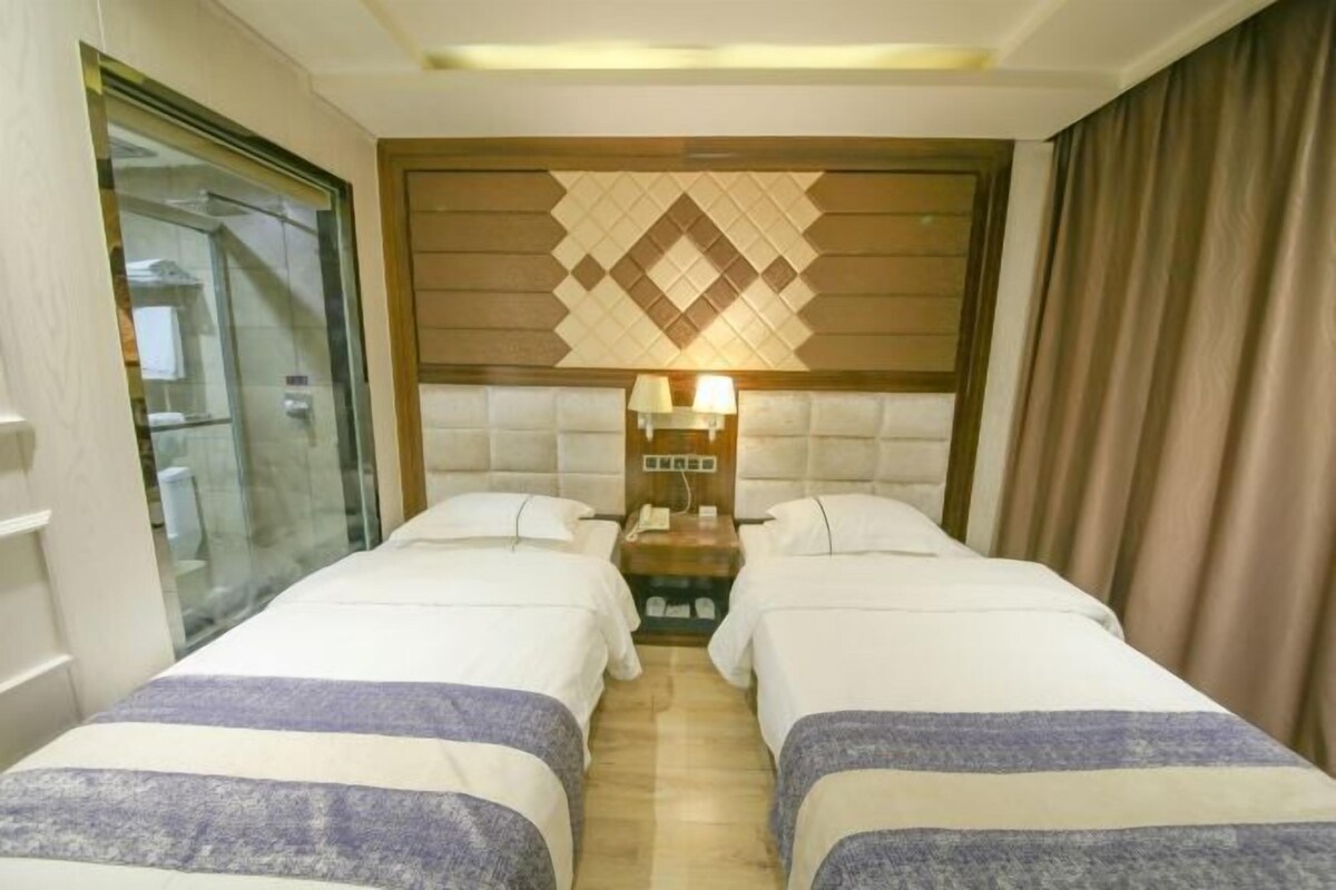 Une chambre d’hôtel avec deux lits, une table de chevet avec des lampes, une tête de lit à motifs et une porte en verre donnant sur la salle de bain.