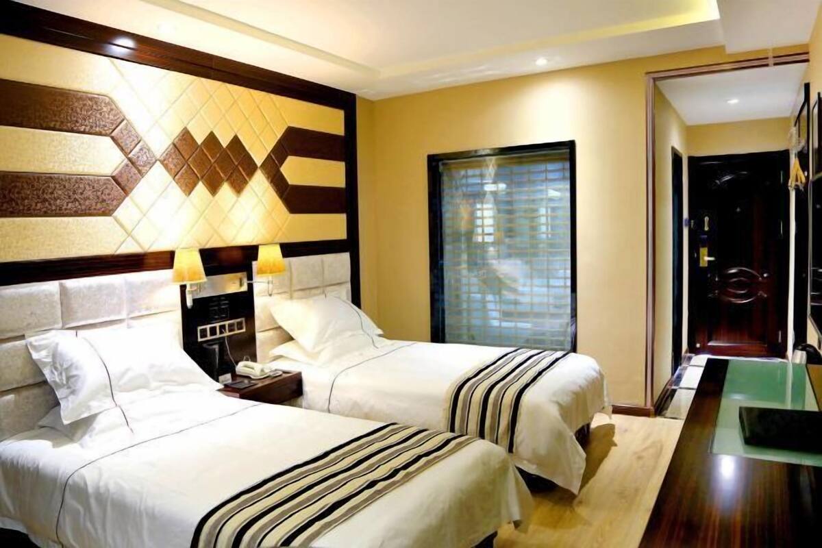 Habitació d'hotel amb dos llits, una tauleta de nit, una làmpada, una finestra amb persianes i una porta.