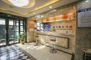 A hotel reception area with a marble counter, two bar stools, a potted plant, and a chandelier.