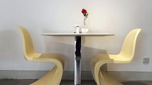 A small round table with two yellow chairs, a red rose in a clear vase, and a small white object on the table.