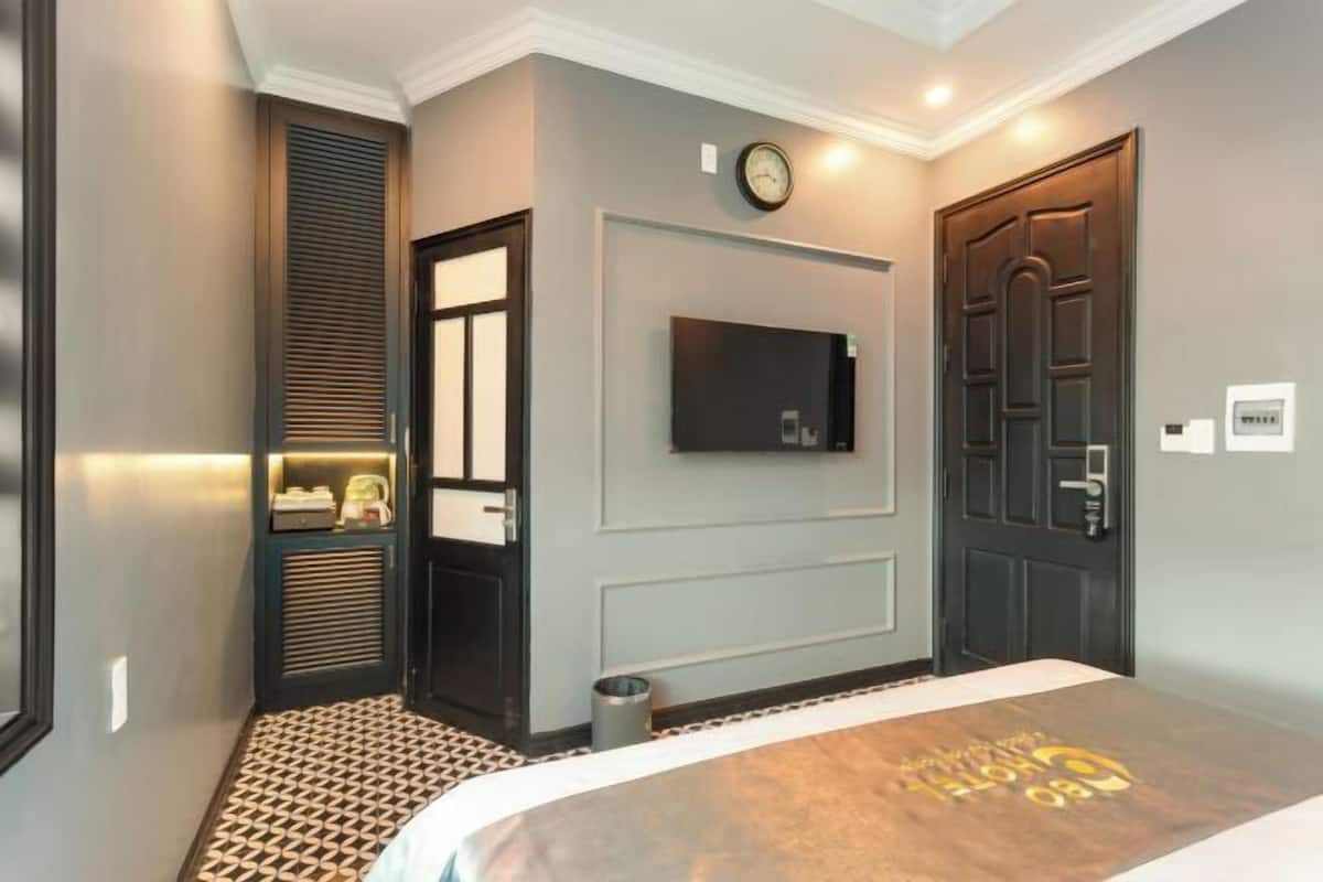 Una habitación de hotel con cama, televisor montado en la pared, una puerta y un cubo de basura pequeño.