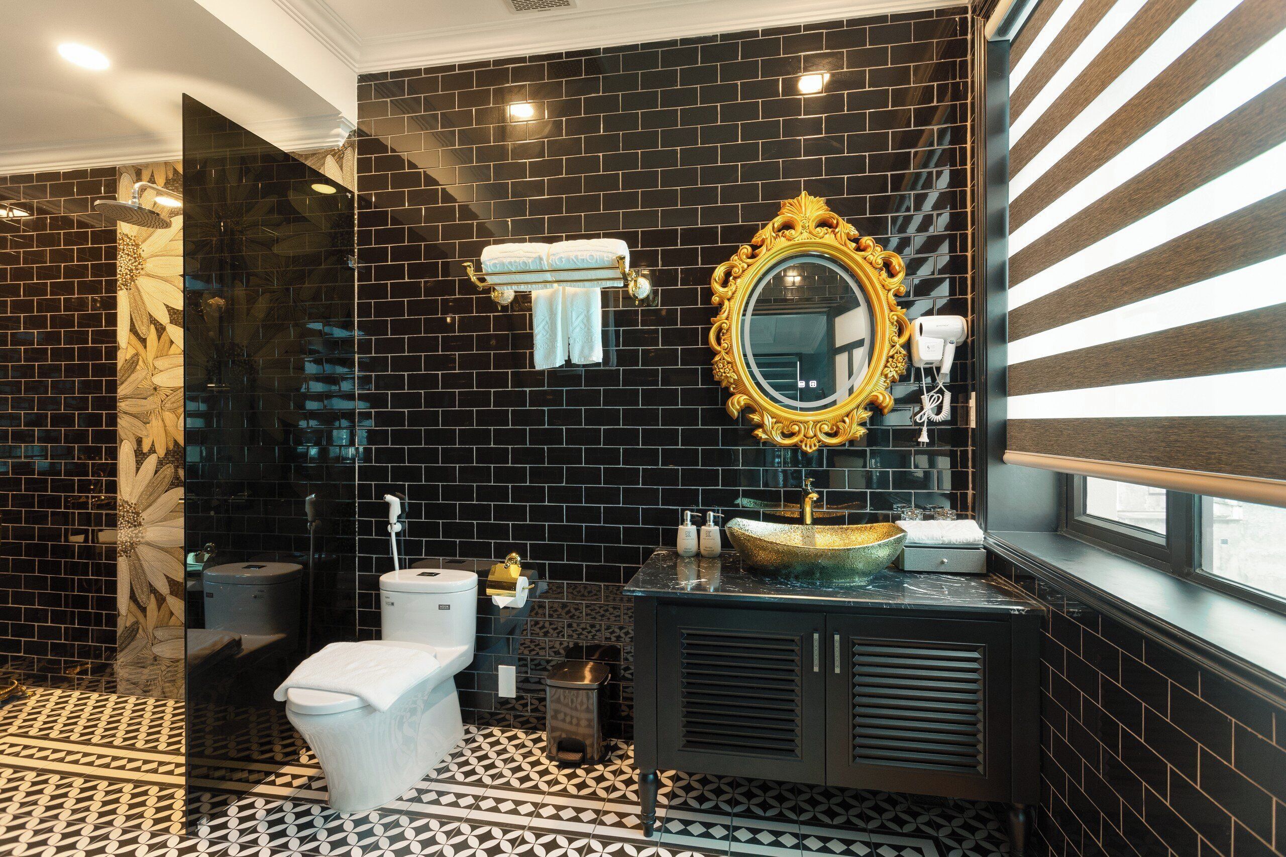 Un baño con baldosas de diseño blanco y negro, un espejo con marco dorado, un inodoro blanco y un lavabo con recipiente dorado.