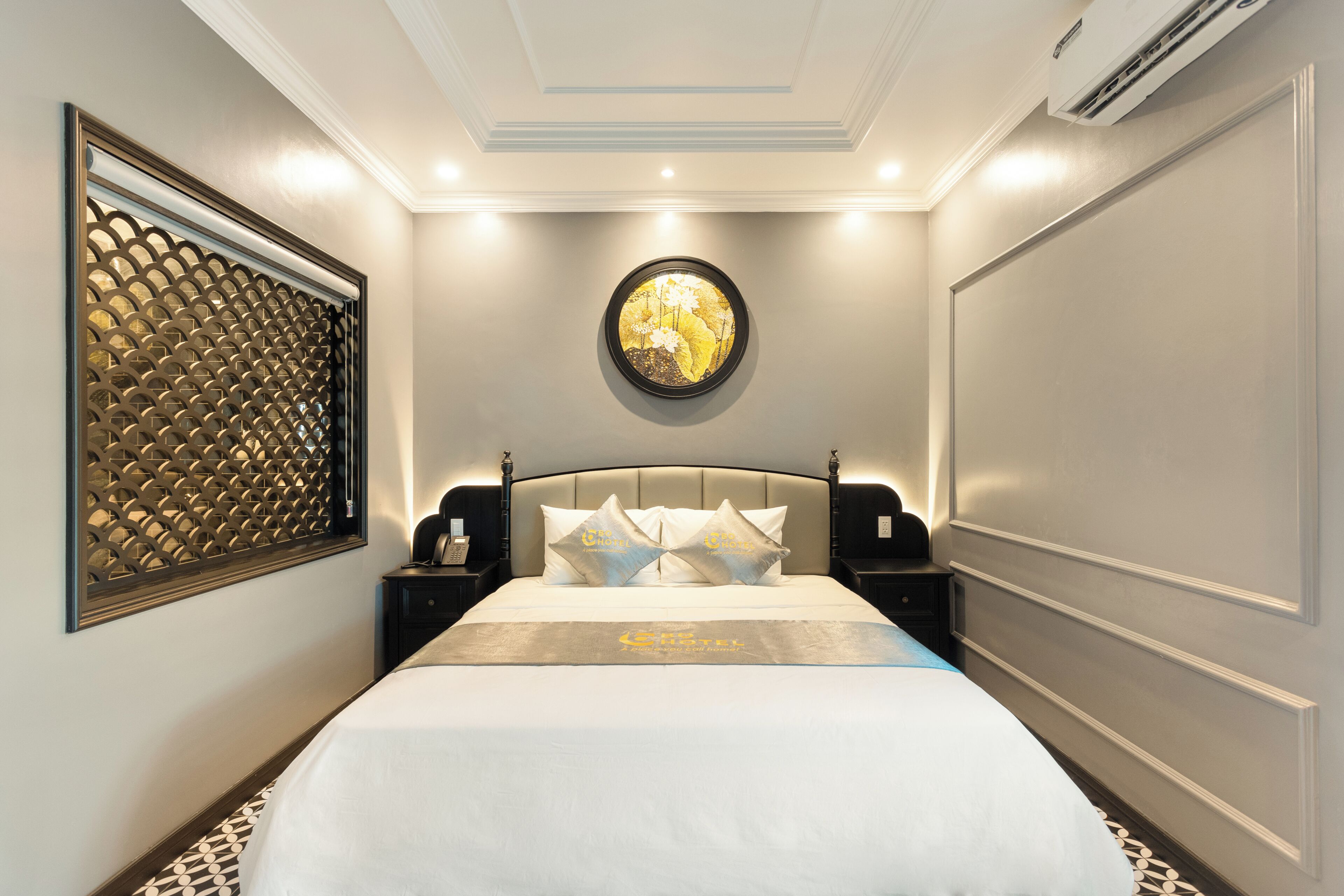 A hotel room with a large bed, a decorative wall panel, a round mirror, and a nightstand with a lamp.