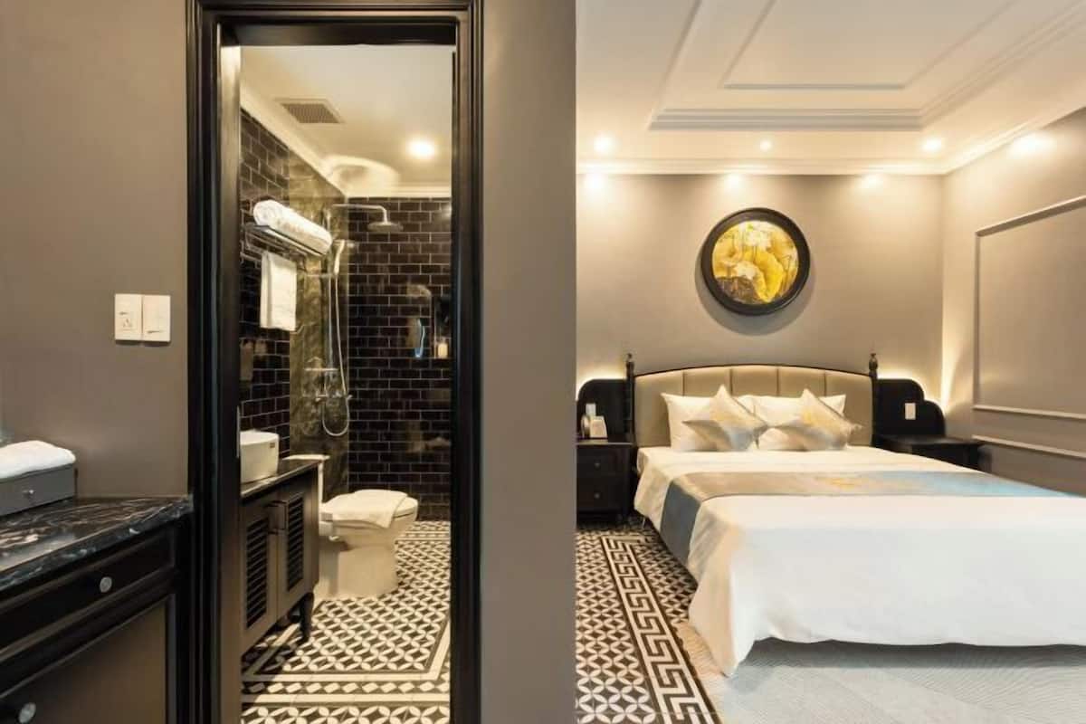 Habitación de hotel con baño, una cama con ropa de cama blanca, un espejo redondo con un cuadro y un piso estampado.