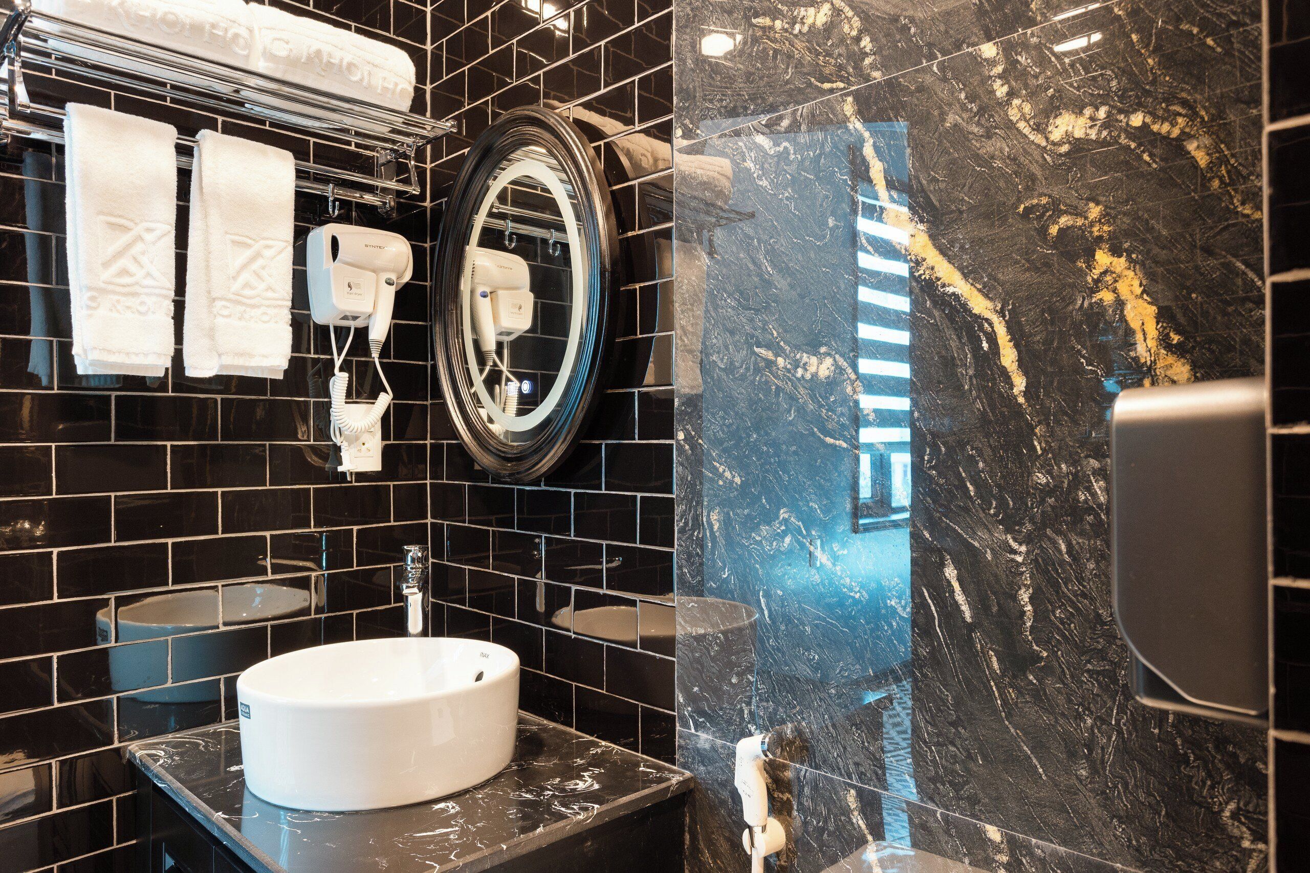 Un baño con pared de azulejos negros, un lavamanos blanco, un espejo y un toallero con toallas.