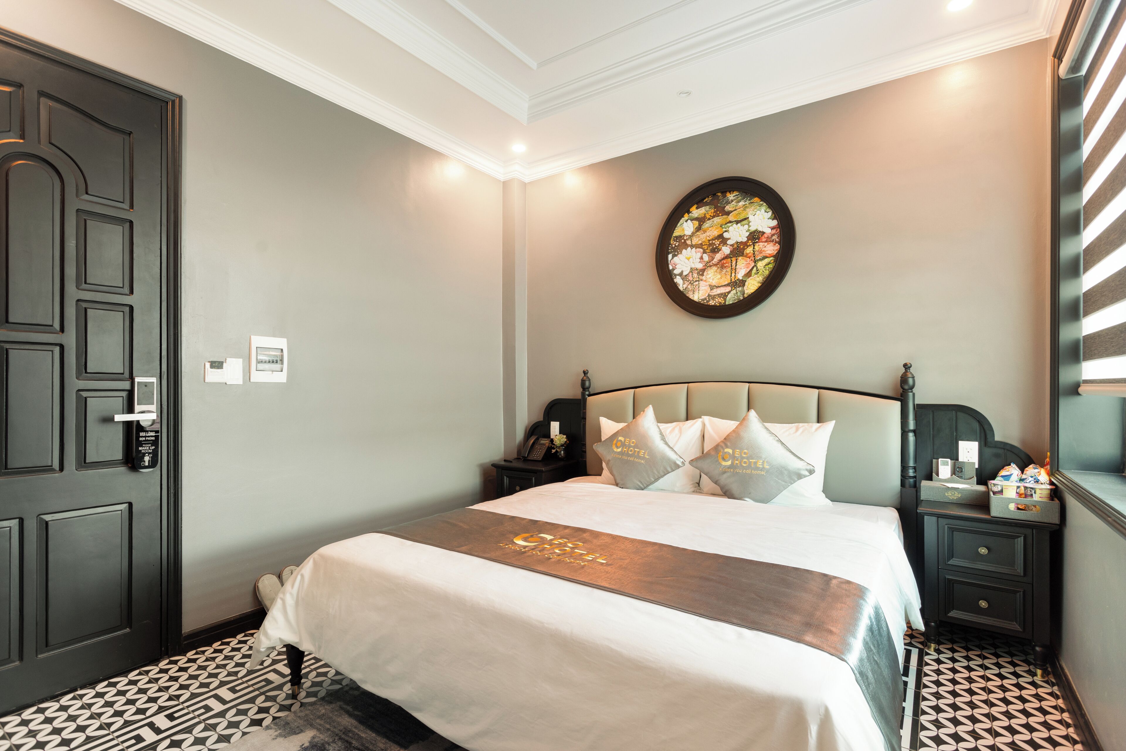 A hotel room with a bed, a nightstand, a wall clock, a painting, and a patterned floor.