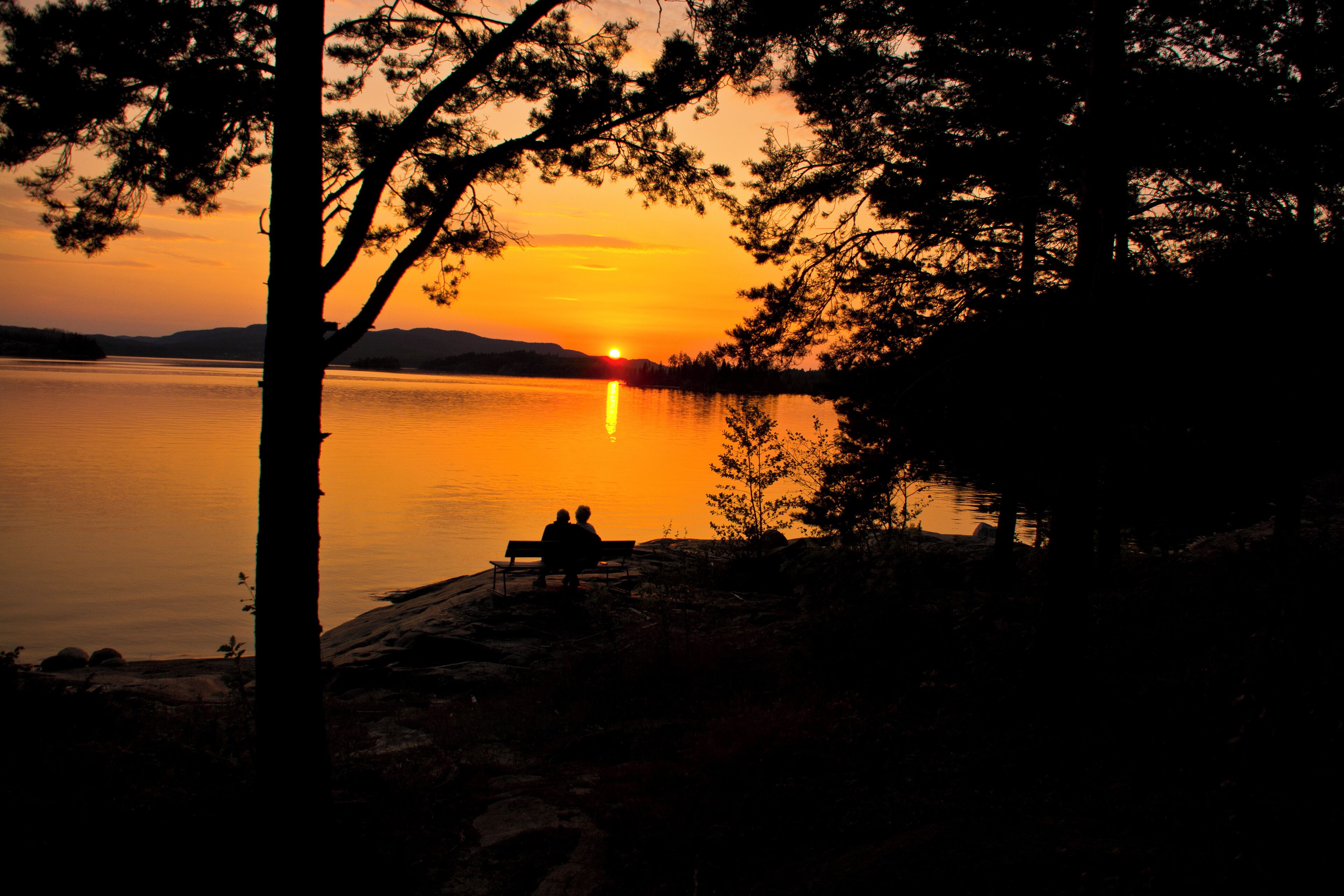 A sunset over a calm lake with a bench and two people sitting on it.