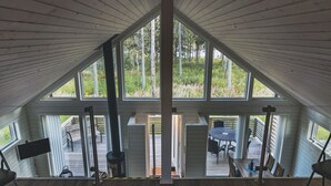 Korkeakattoinen huone, jossa on suuret ikkunat, ulkona puinen terassi sekä pöytä ja tuolit.