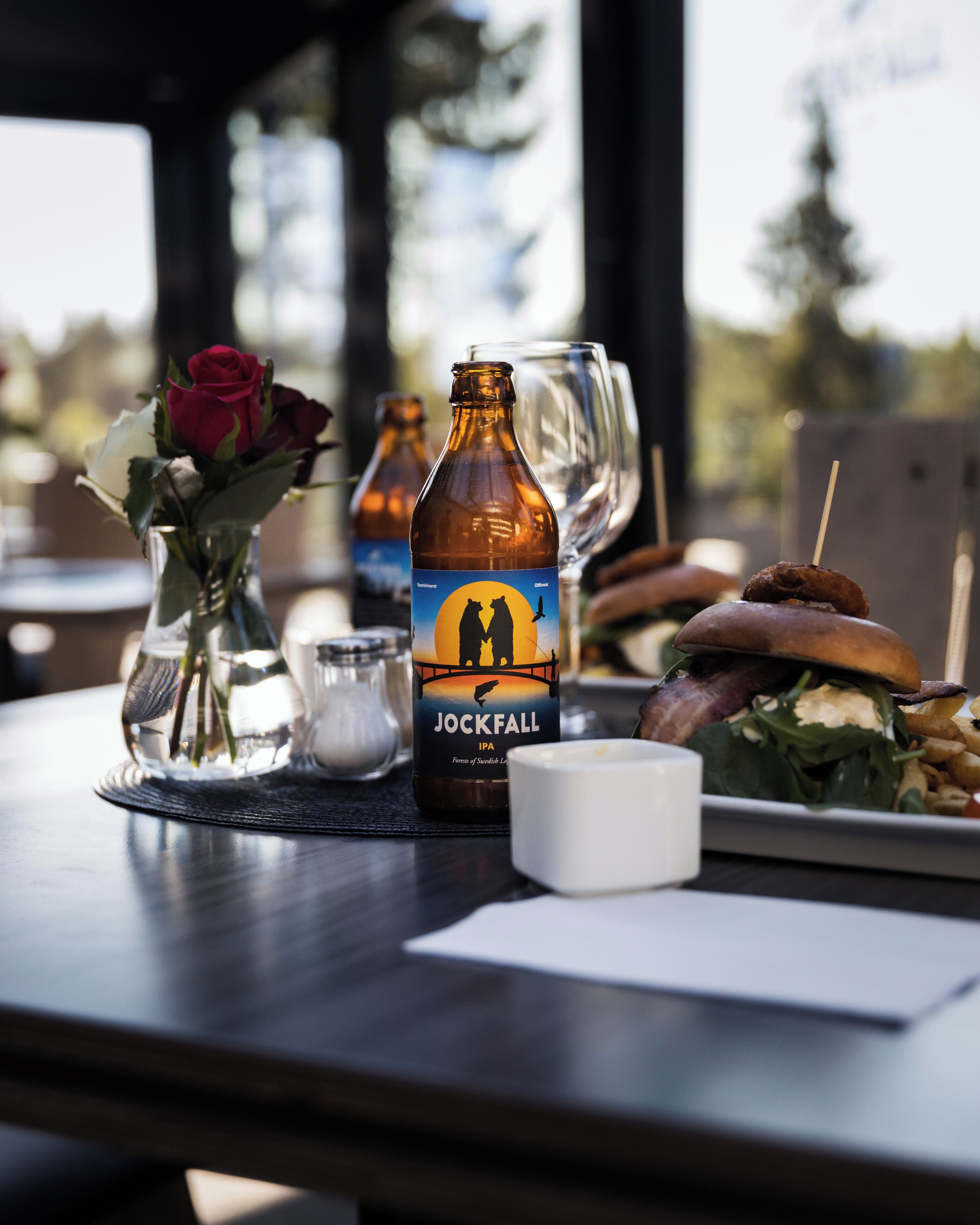 A table with a bottle of Jockfall IPA, a candle, a glass of water, a plate with food, and a vase with flowers.
