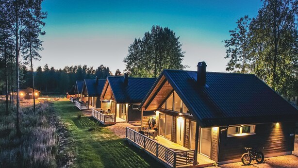 Rivi puisia mökkejä, joissa on suuret ikkunat, sijaitsee metsäisellä alueella. Mökkien eteen johtaa sorapolkua.
