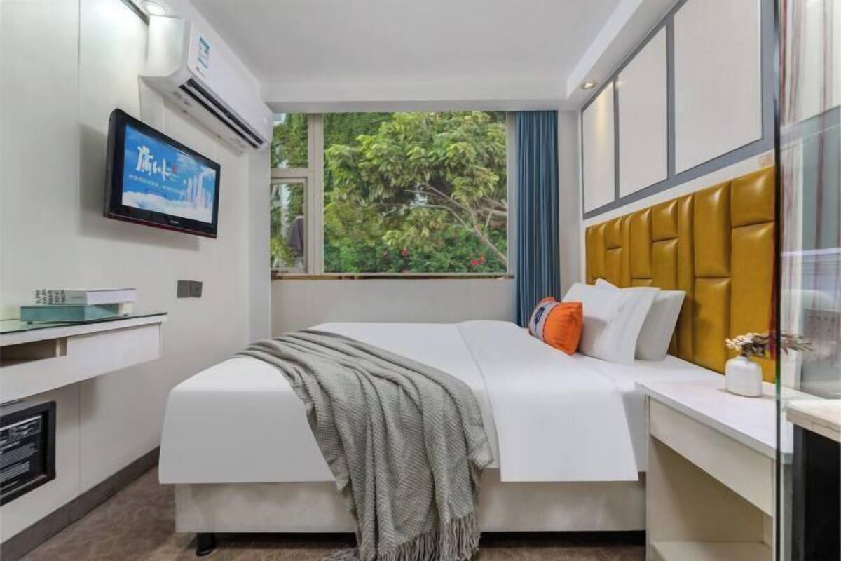 A modern hotel room with a bed, a TV, a window with a view of greenery, and a small desk with a computer.