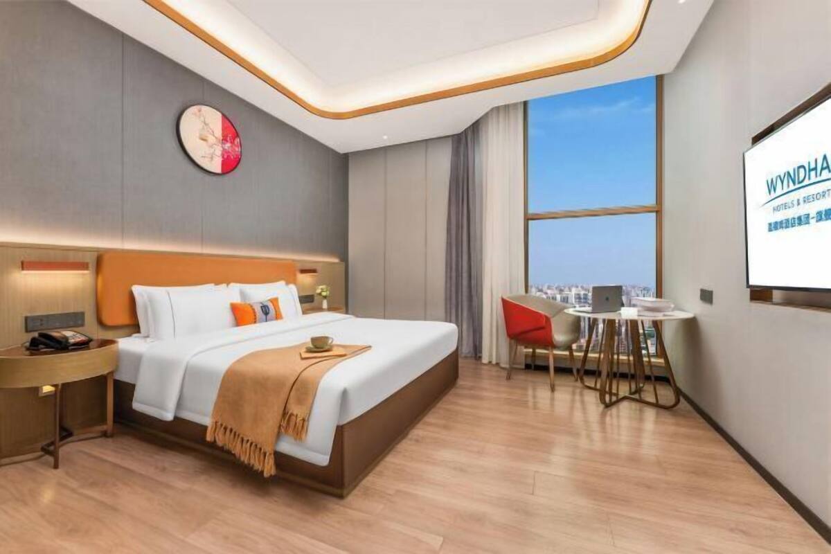 A hotel room with a bed, bedside table, desk, chair, and a large window with a city view.