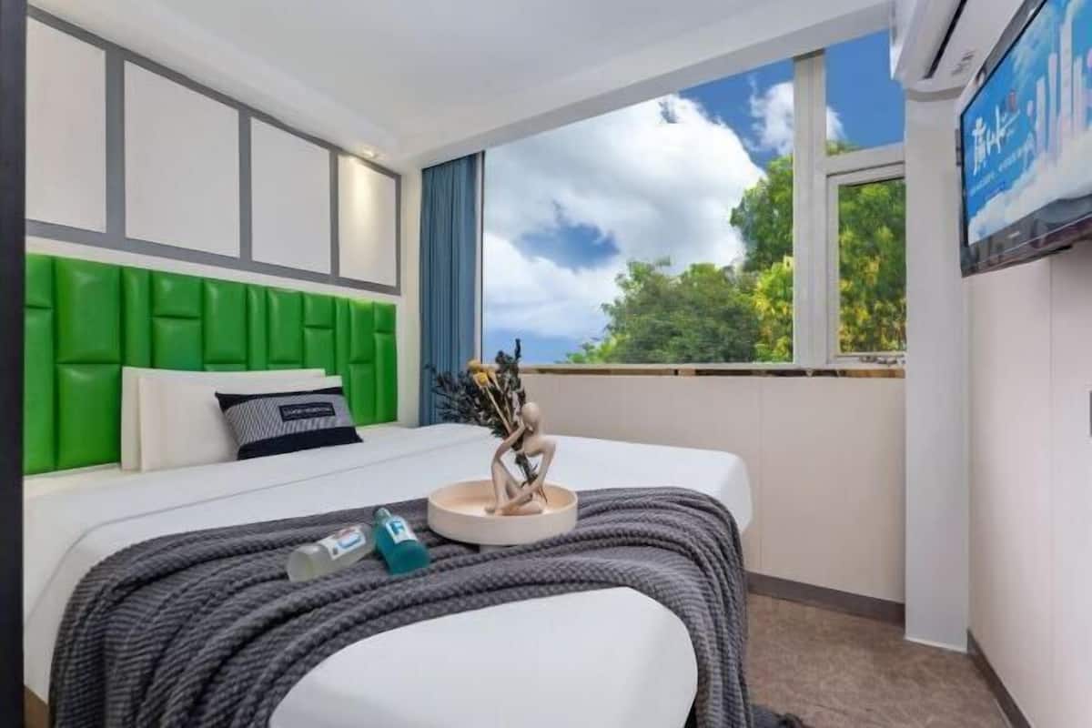 A hotel room with a bed, a green headboard, a window with a view of trees, and a television on the wall.