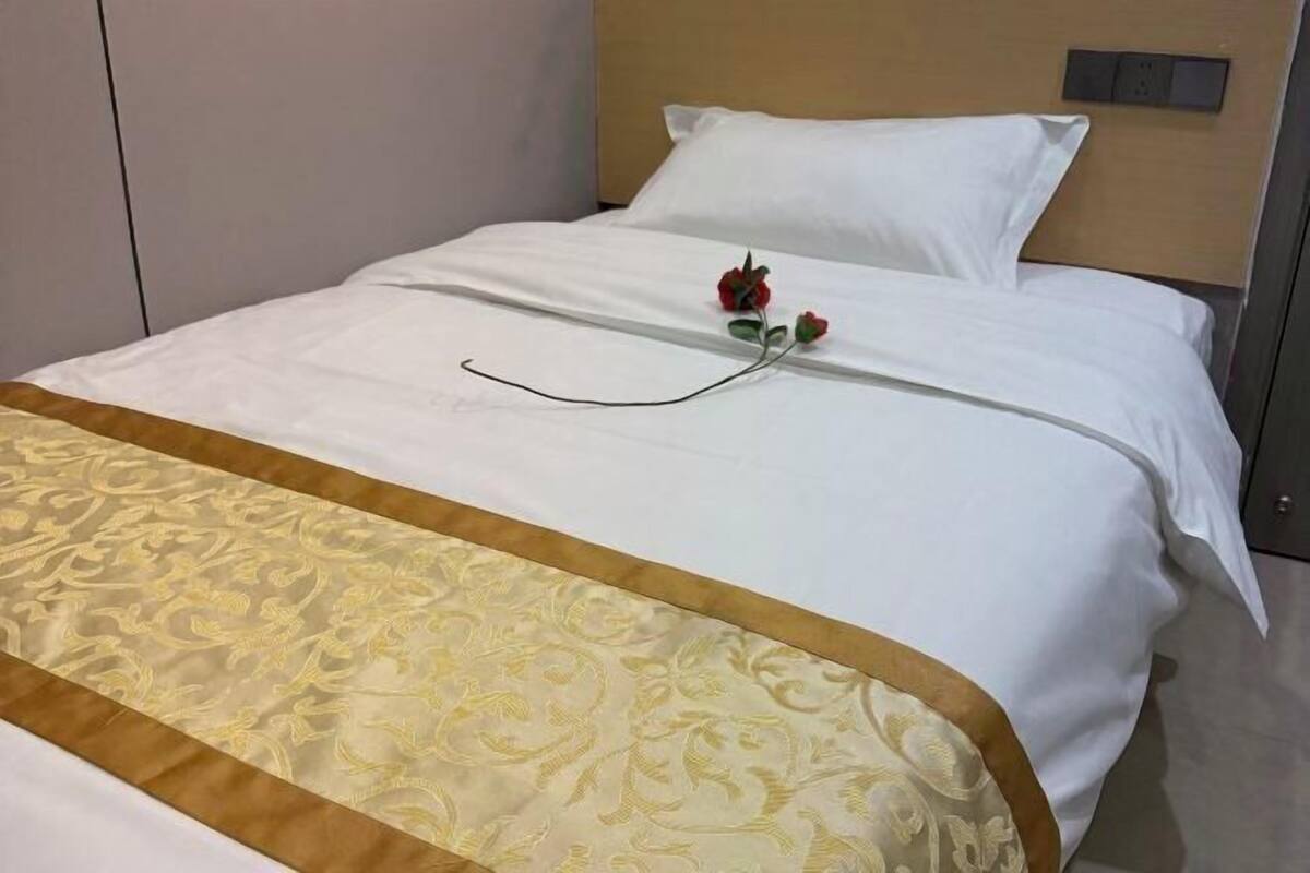 A neatly made bed with a white sheet, a decorative golden bed runner, and a single red rose on the bed.