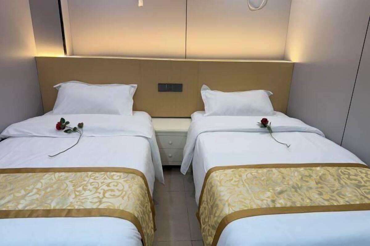 A hotel room with two beds, each with a white bedspread and a decorative gold trim. There are bedside tables with lamps and a wall-mounted control panel.