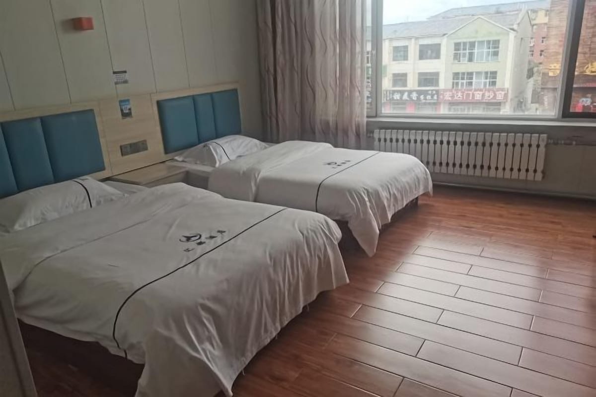Een hotelkamer met twee bedden, een raam met gordijnen, een radiator en uitzicht op gebouwen.