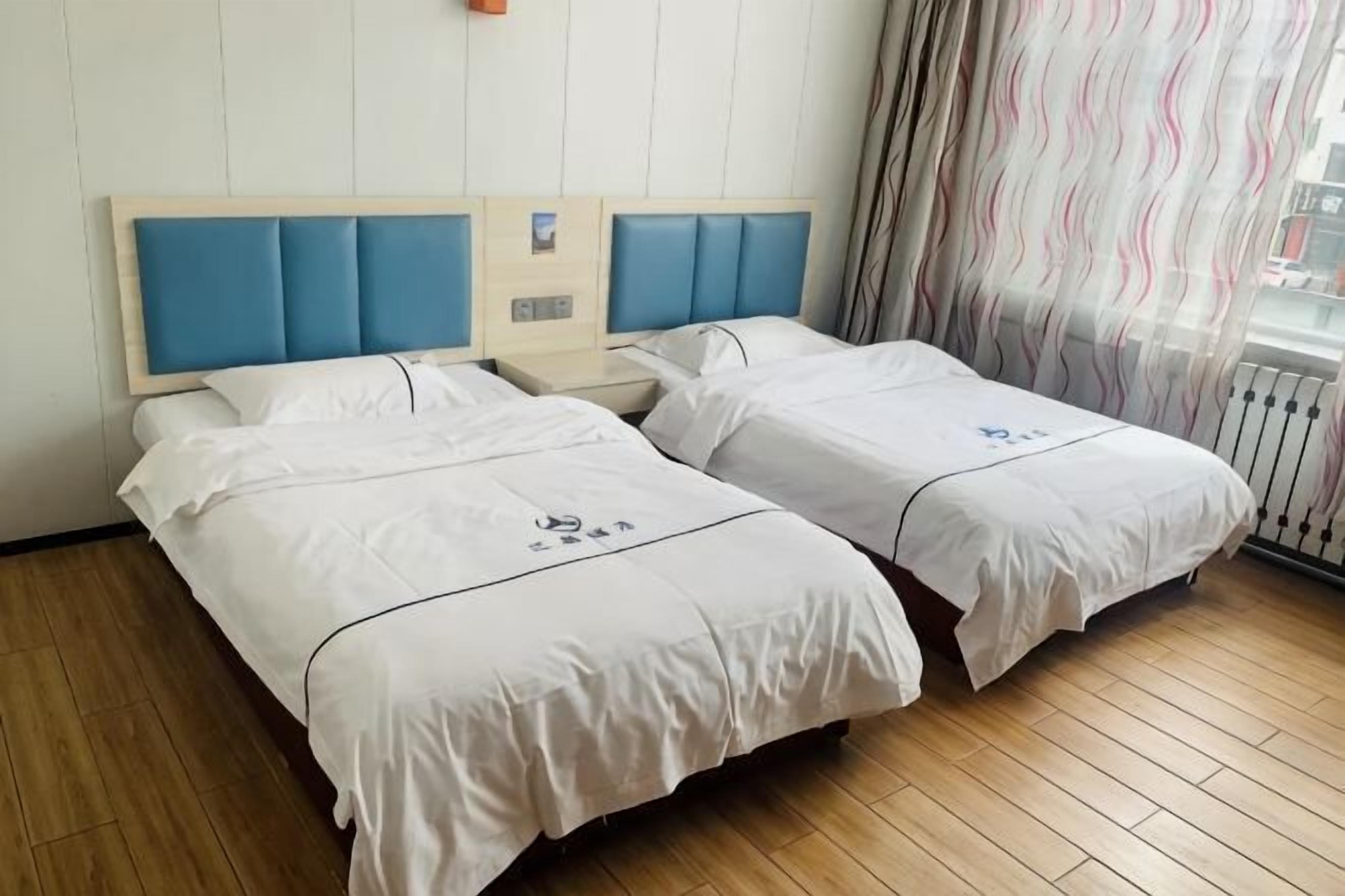 A hotel room with two beds, a wooden floor, a window with curtains, and a white wall.