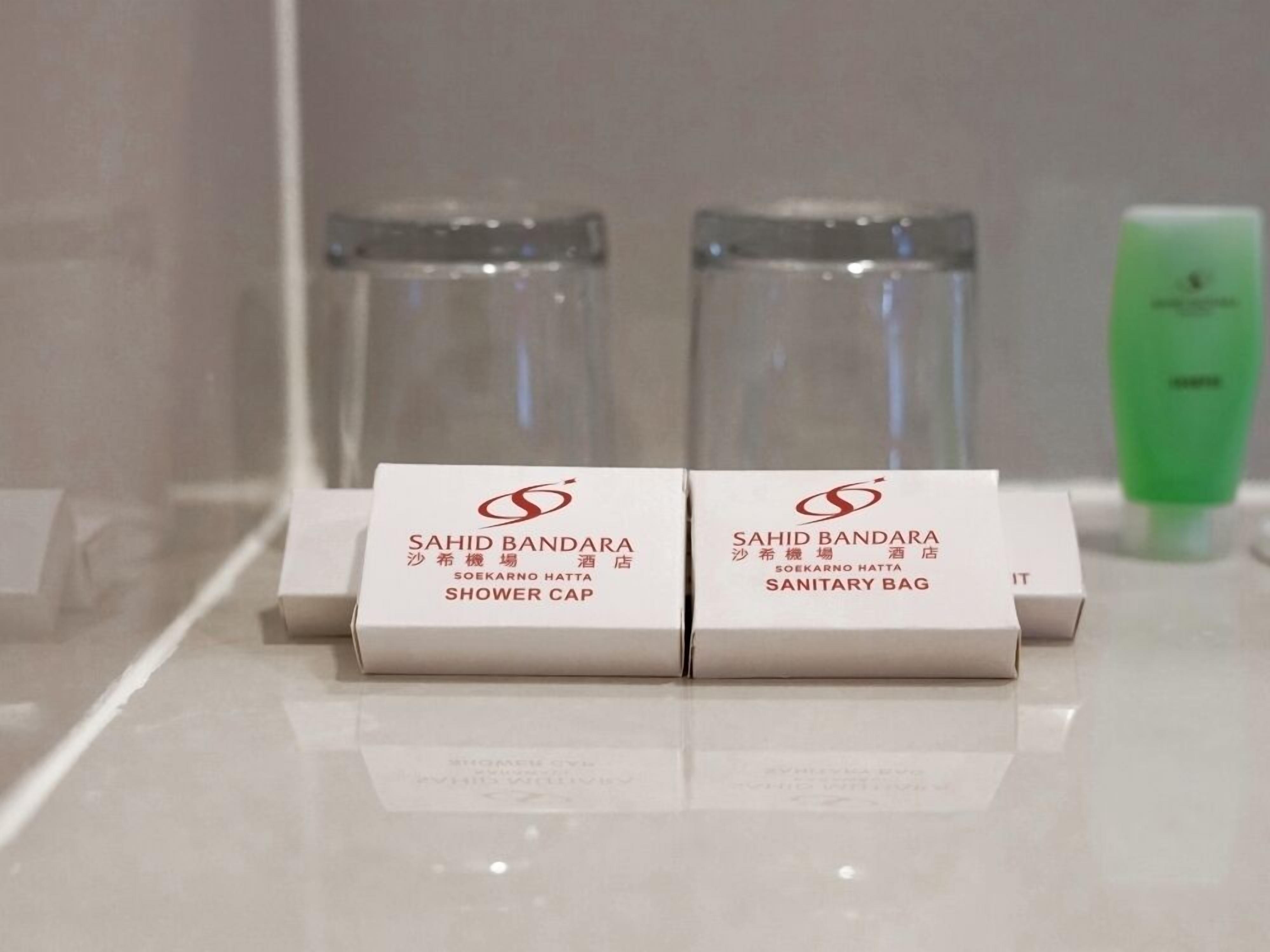Twee dozen met de tekst 'SAHID BANDARA SHOWER CAP' en 'SAHID BANDARA SANITARY BAG' op een reflecterend oppervlak, met twee heldere flessen en een groene fles op de achtergrond.