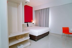 Una habitación de hotel moderna con una cama grande, un armario, una pared de acento rojo brillante, una ventana con cortinas y una sola silla naranja.