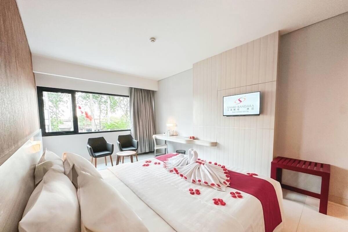 A hotel room with a bed, two chairs, a desk, a TV, and a window with curtains.