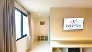 Une chambre d’hôtel avec une grande fenêtre, un panneau fixé au mur, un tableau encadré et une étagère avec un panier et des chaussons.