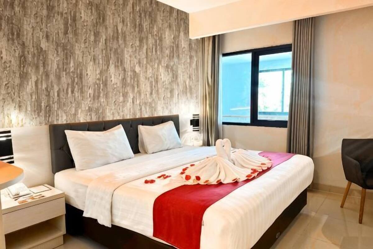 A hotel room with a large bed, a nightstand, a chair, and a view of the outdoors through a window.