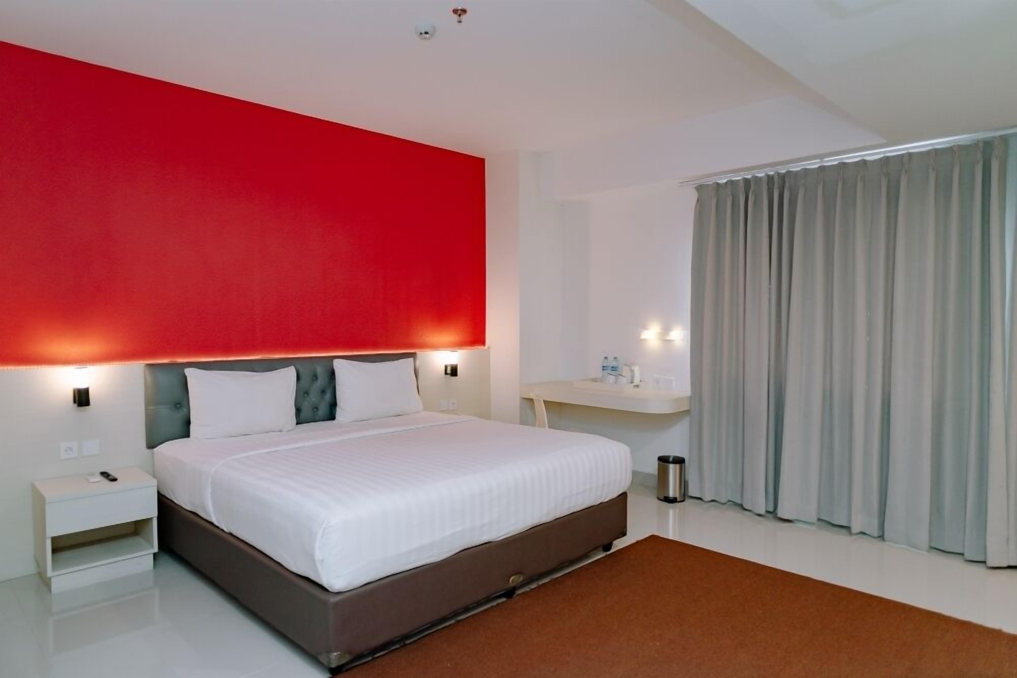 A modern hotel room with a large bed, a nightstand, a desk with a chair, and a window with curtains.