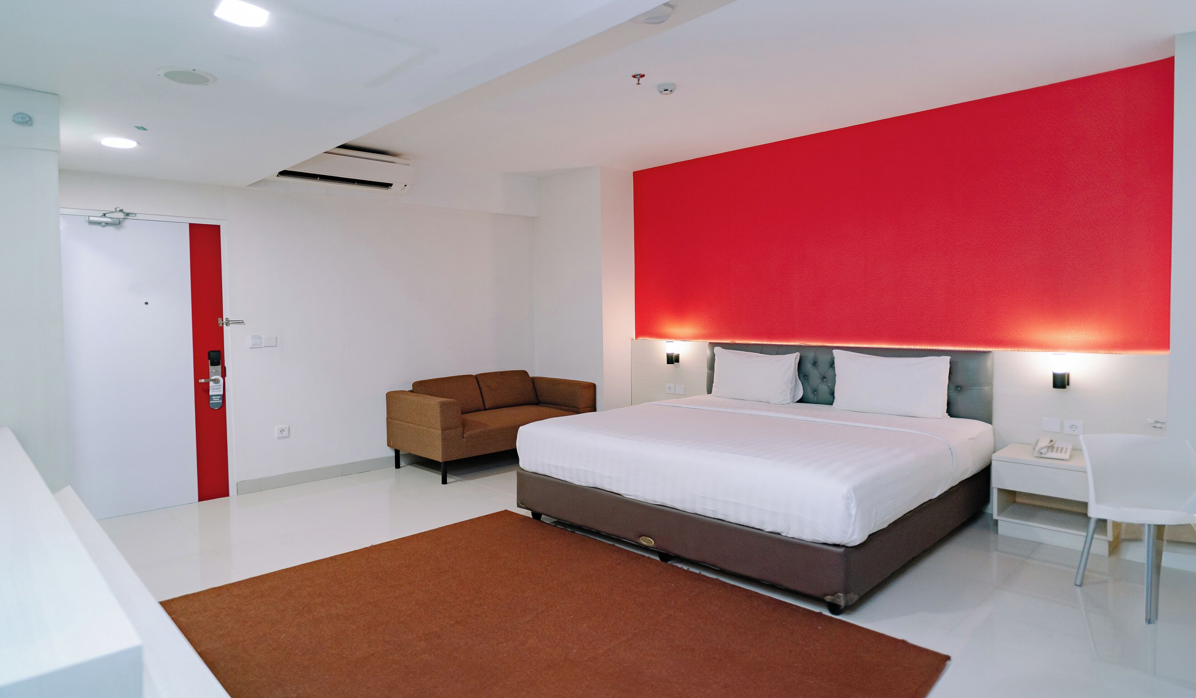 A modern hotel room with a large bed, a red accent wall, a brown sofa, and a white desk.