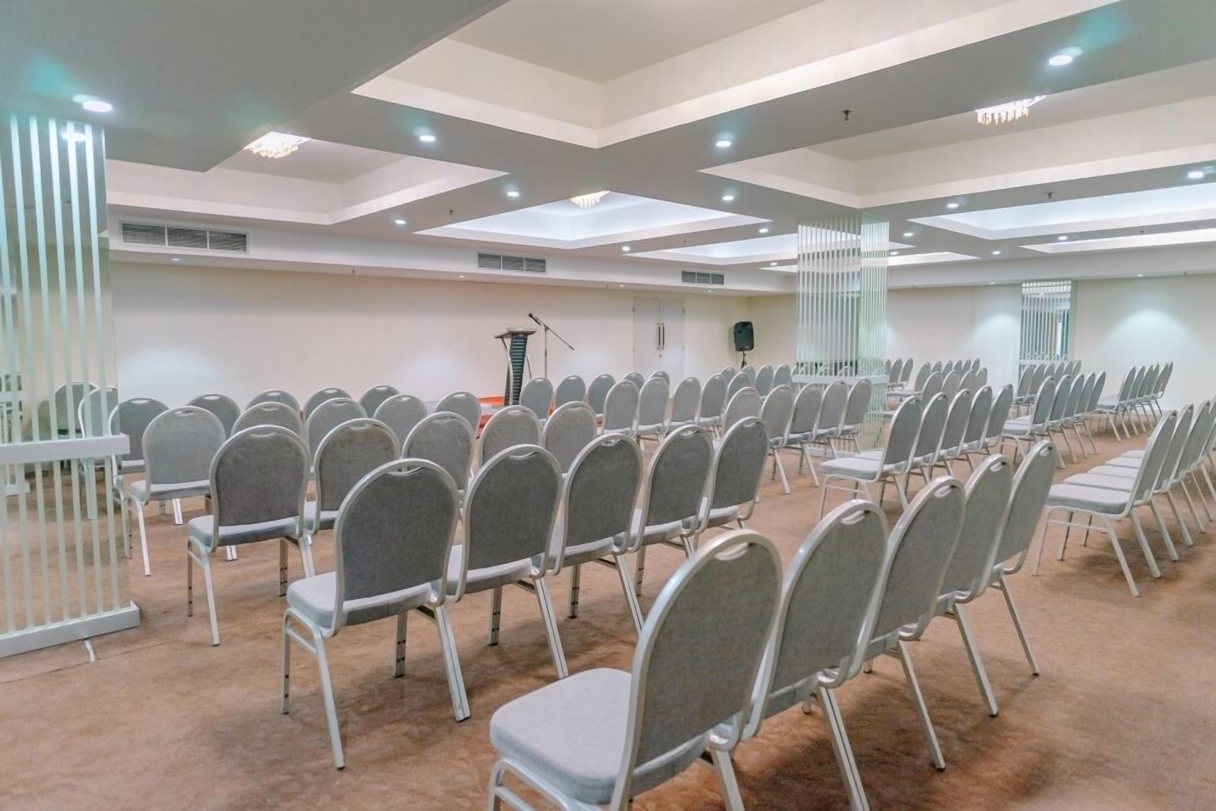 Un salón de conferencias con filas de sillas grises, un atril y un orador a la derecha.