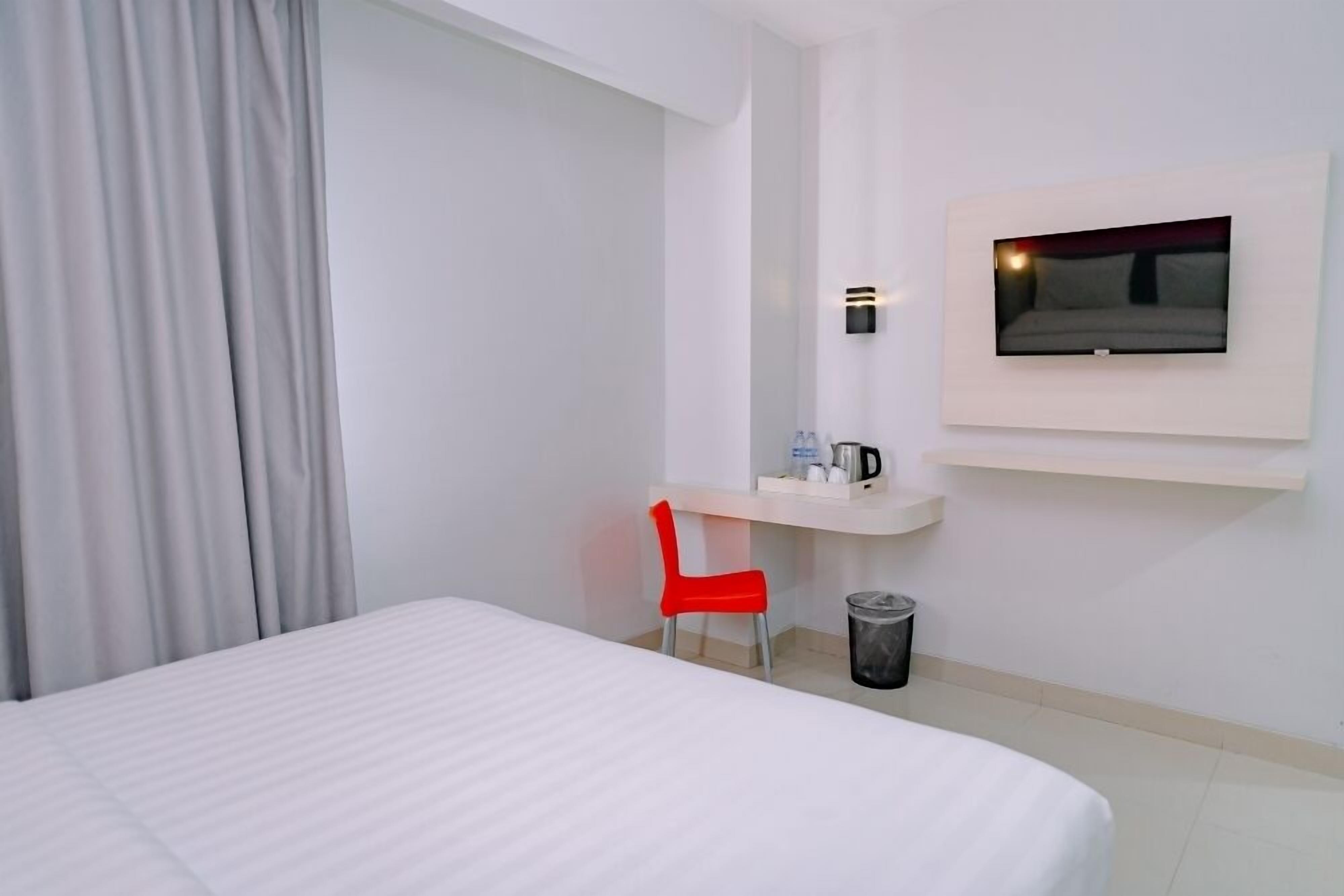 A hotel room with a bed, a red chair, a desk with a TV mounted above it, and a trash can.