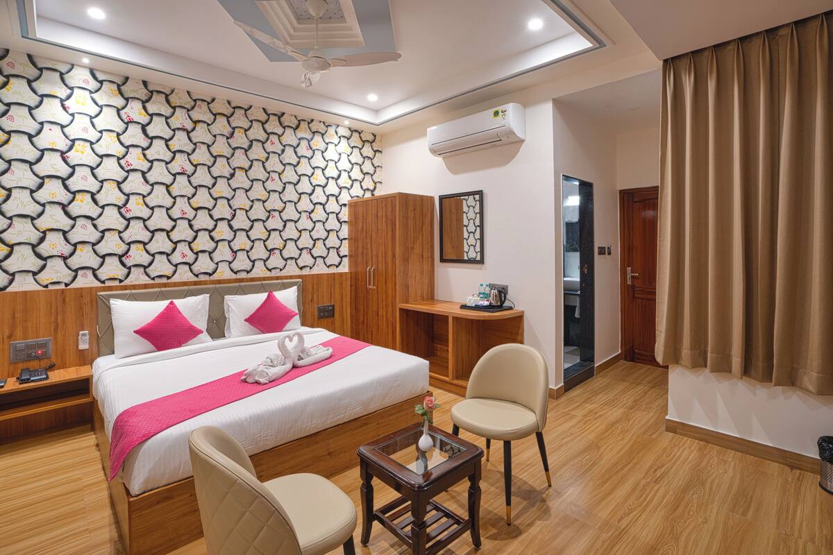 A hotel room with a bed, two chairs, a small table, a wardrobe, a mirror, and an air conditioning unit.