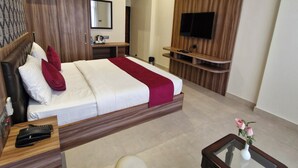 A modern hotel room with a large bed, a flat-screen TV mounted on a wooden panel, a glass coffee table, a white armchair, and an air conditioning unit. - Hotel Aranyasutra (Udaipur)