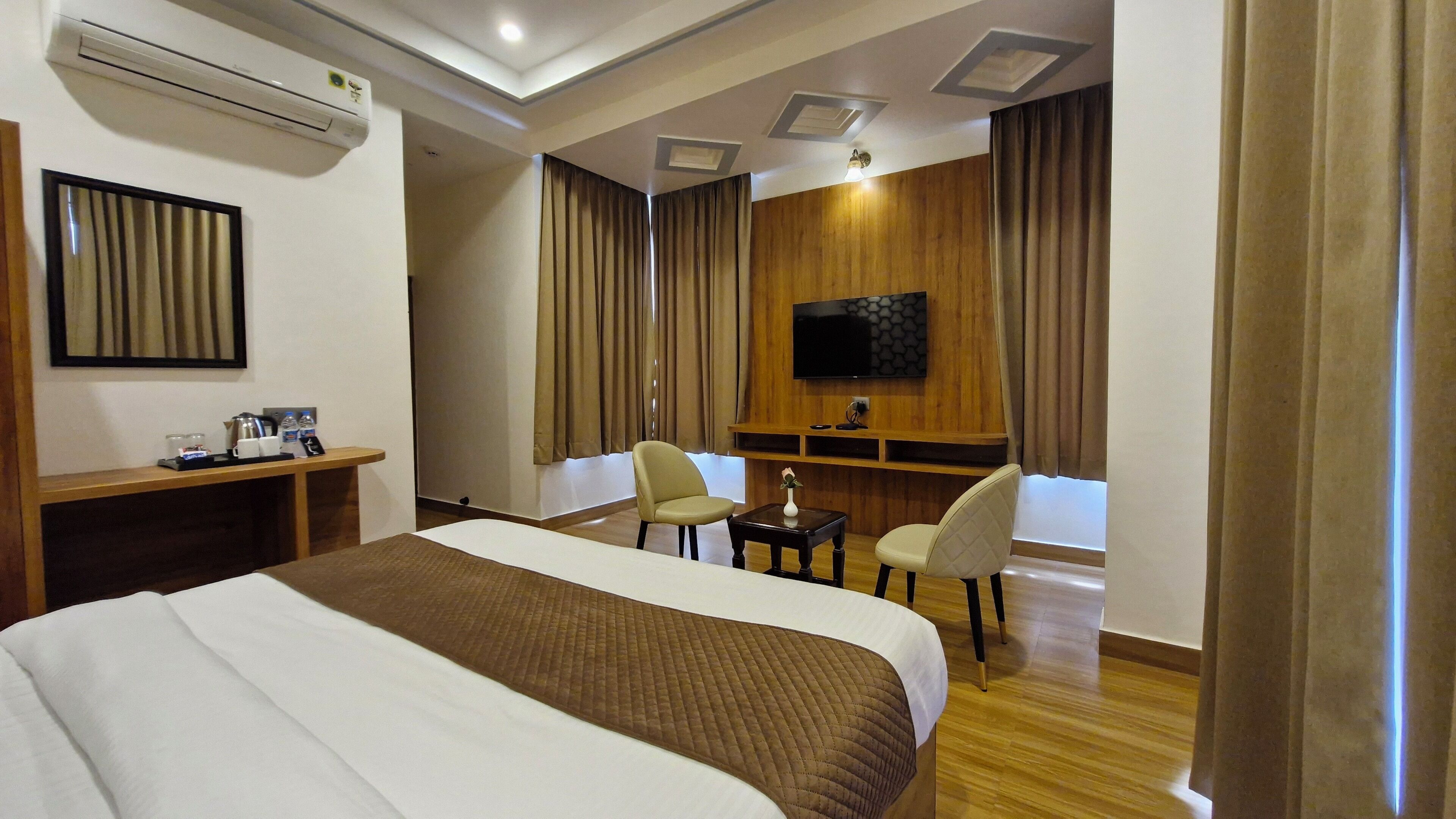 A hotel room with a large bed, a desk with a chair, a small table with a vase, and a TV mounted on the wall.