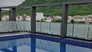 A rooftop swimming pool with clear blue water, surrounded by a tiled deck and metal railings. In the background, there is a lush green hill with a castle-like structure on top. - Hotel Aranyasutra (Udaipur)