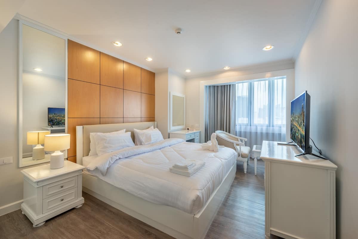 A modern hotel room with a large bed, a desk, a chair, a TV, and a large window with curtains.