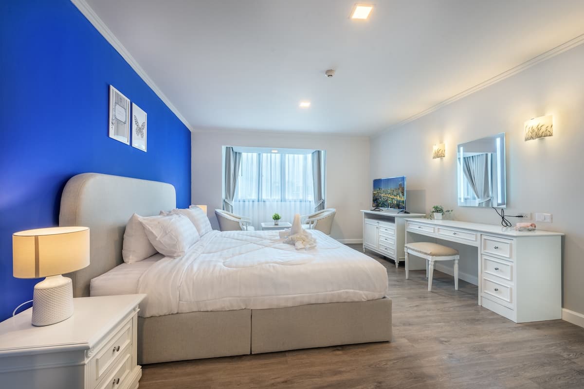 A bedroom with a bed, bedside table, lamp, desk, chair, and a large window with curtains.