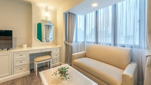A modern hotel room with a large window, a sofa, a desk with drawers, a bench, and a television. - Neom Town Residence (Bangkok)