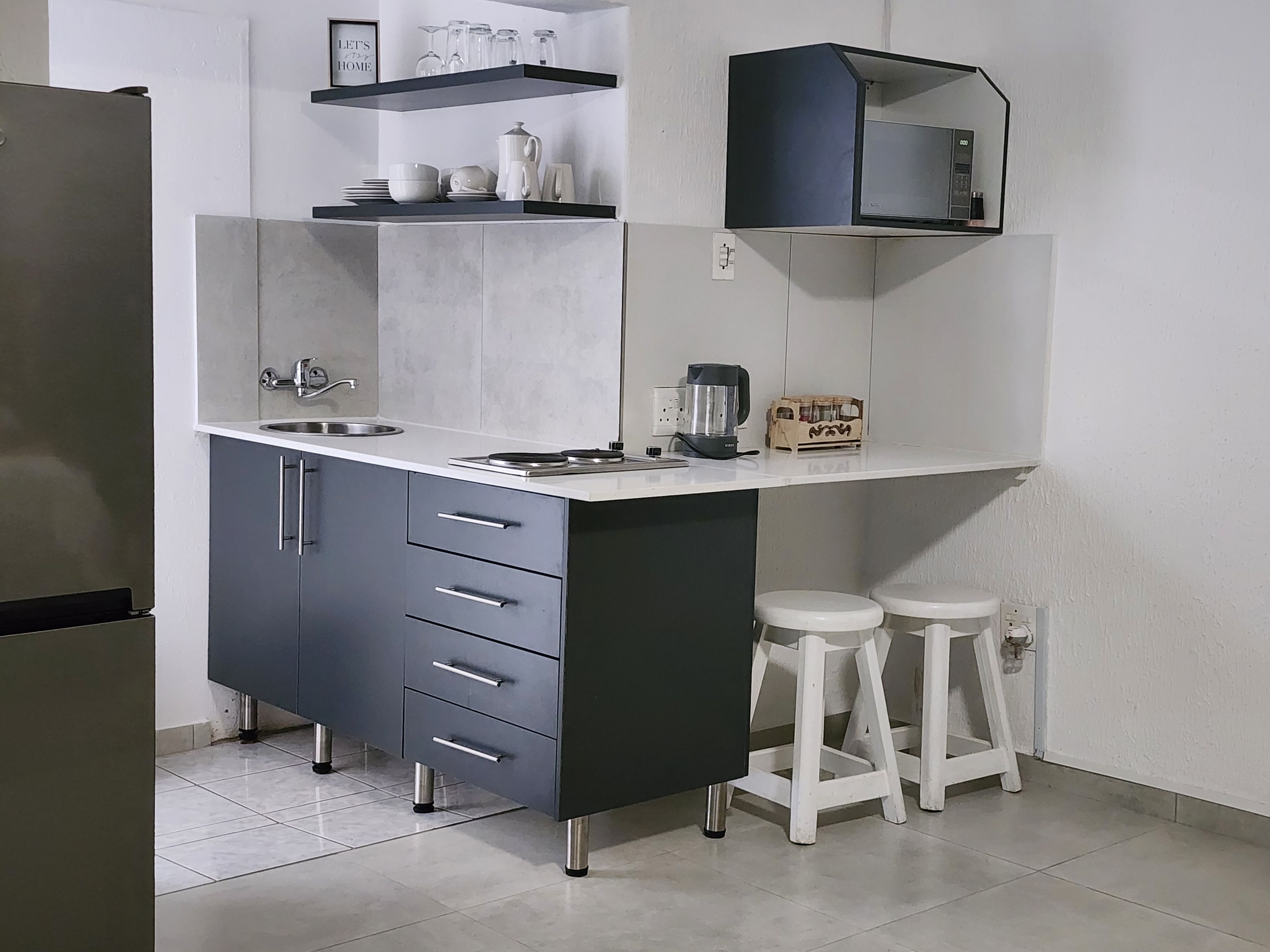 Cocina compacta con pileta integrada, cafetera y microondas. Hay dos taburetes y una estantería con utensilios de cocina.