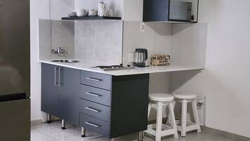 Cocina compacta con pileta integrada, cafetera y microondas. Hay dos taburetes y una estantería con utensilios de cocina.