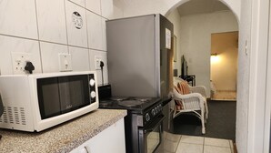 A compact kitchen with a microwave, stove, and oven. There is a white cabinet with handles, a chair, and a small table with a plant. - Room-busa House - Triple Room (Mbombela)
