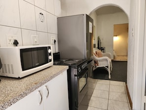 A compact kitchen with a microwave, stove, and oven. There is a white cabinet with handles, a chair, and a small table with a plant.