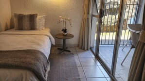 A bedroom with a bed, a small table with a vase, a window with curtains, and a view of the outside. - Room-busa House - Triple Room (Mbombela)