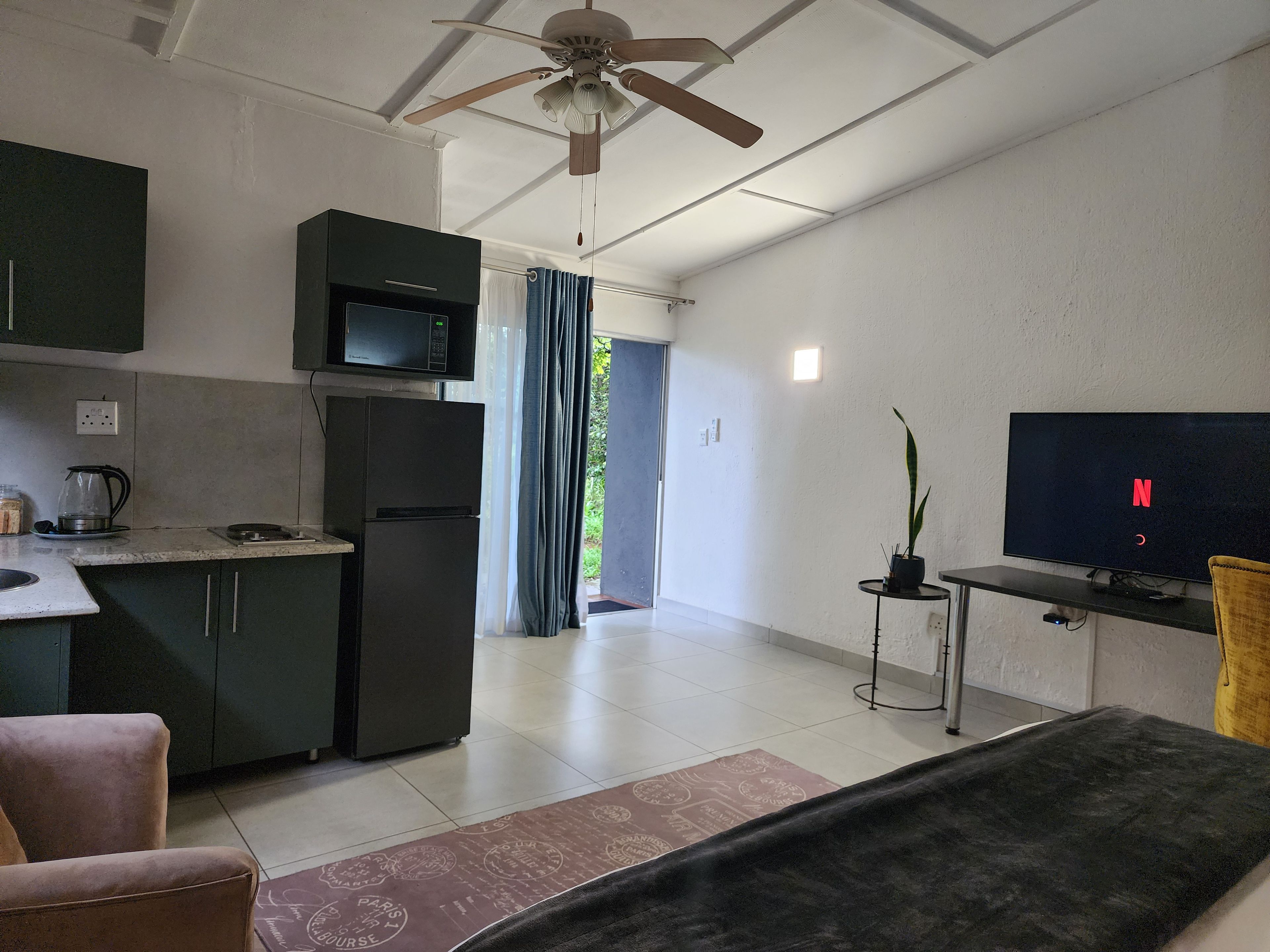 A modern living room with a flat-screen TV, a ceiling fan, and a kitchen area with a microwave and refrigerator.