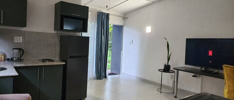 A modern living room with a flat-screen TV, a ceiling fan, and a kitchen area with a microwave and refrigerator.