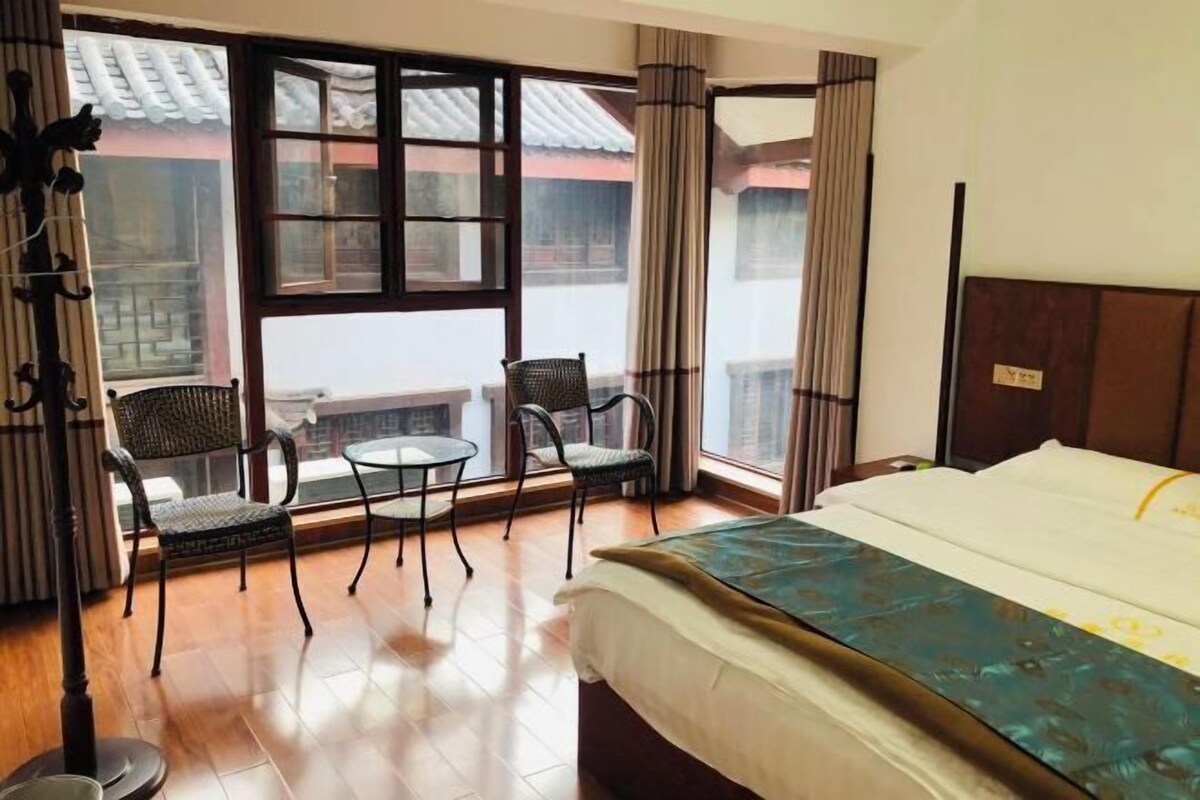 Una habitación de hotel con una cama, dos sillas, una mesita y ventanas amplias.