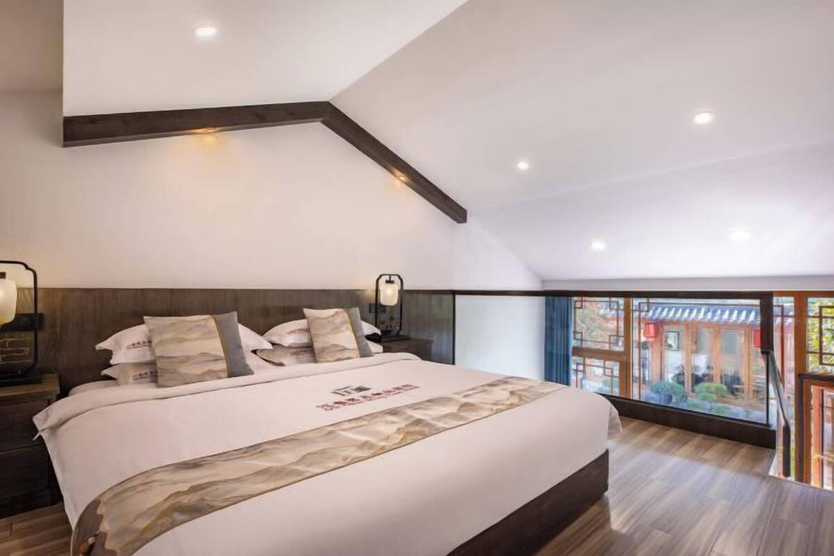Un dormitorio moderno con una cama grande, cabecera de madera, lámparas de noche y vistas a un edificio tradicional a través de ventanas amplias.