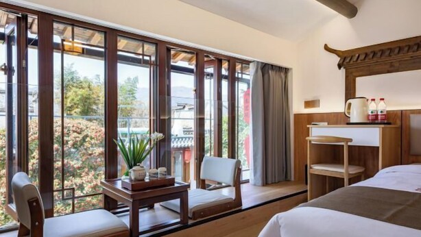 A hotel room with a large window, a bed, a chair, a small table with a plant, and a shelf with bottles.
